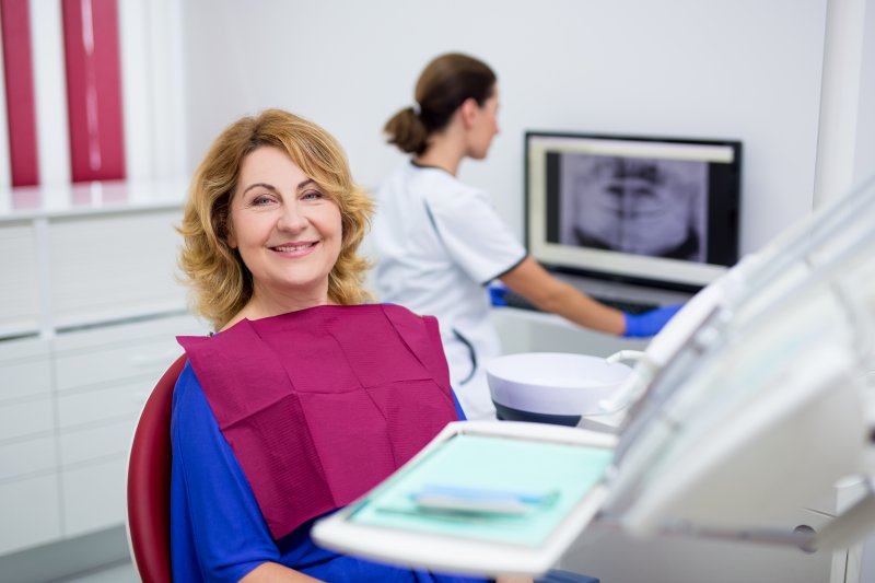 an older woman smiling in the dentist’s chair while the dentist reviews a digital image